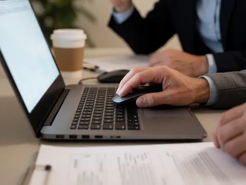 Close-up of a hand using a computer mouse on a laptop, with documents and coffee on the desk for business productivity.