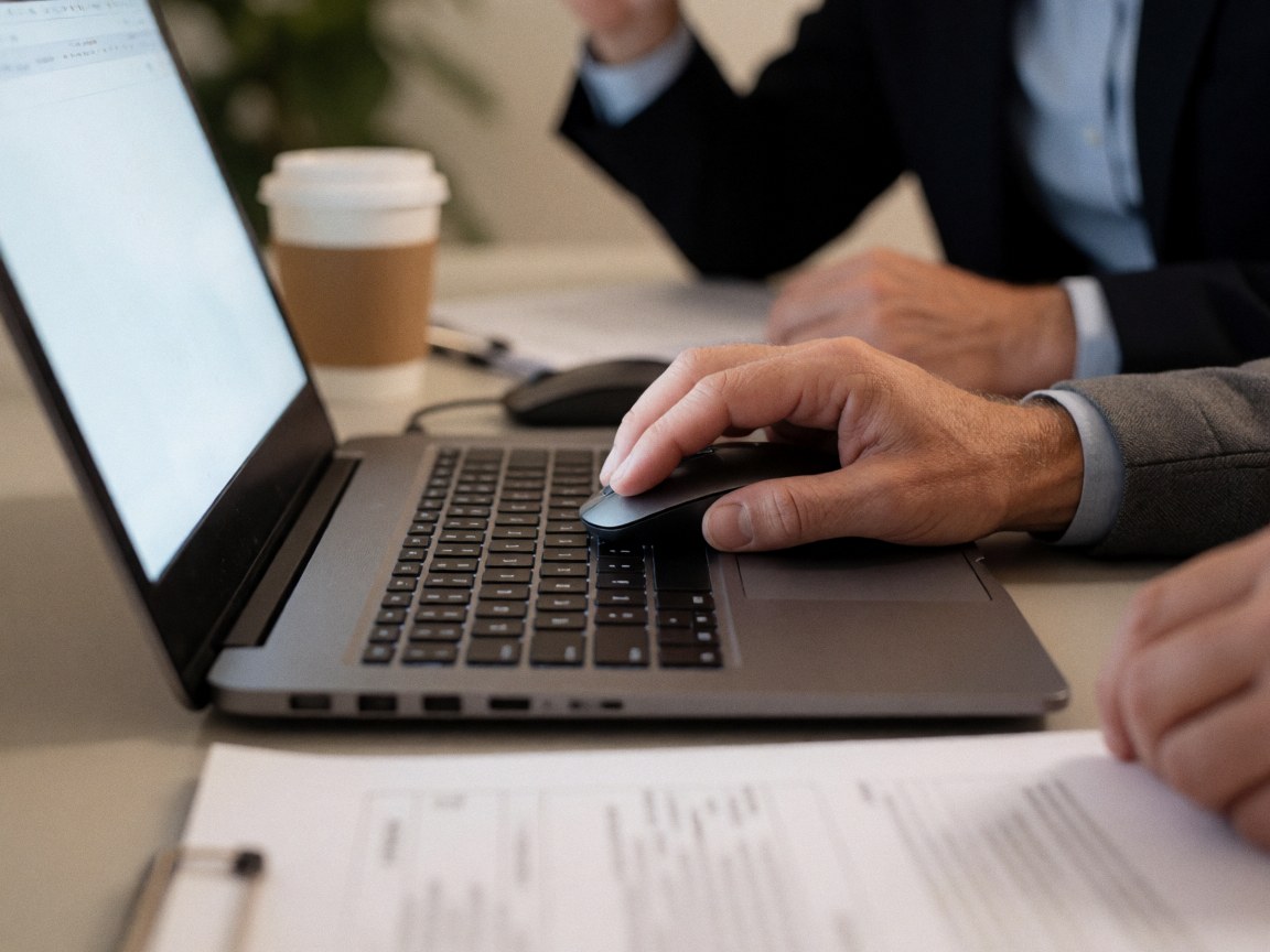 Close-up of a hand using a computer mouse on a laptop, with documents and coffee on the desk for business productivity.