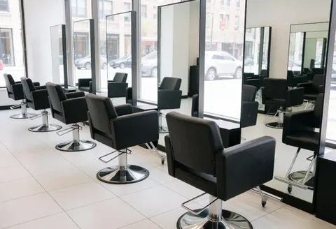 Empty modern hair salon interior featuring black styling chairs, large mirrors, white tiled floors, and natural light from windows