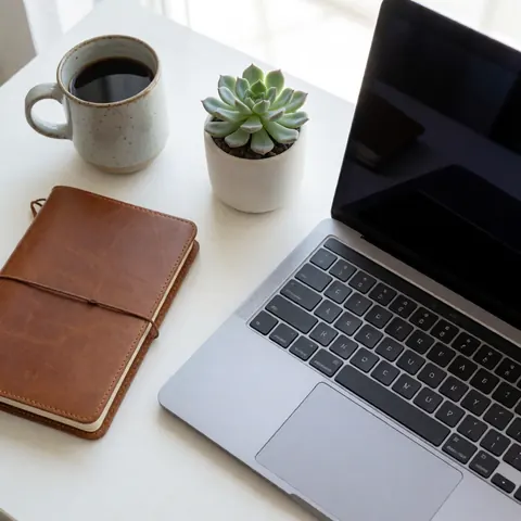 White desk with laptop, leather notebook, coffee cup, and succulent plant under natural light