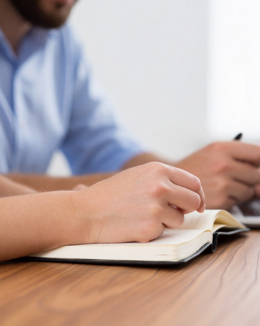 Two people working together at a wooden desk, one reviews a notebook while the other writes with a pen.
