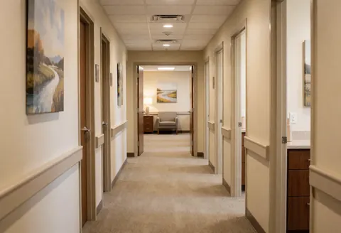 A serene spa corridor featuring soft carpeting, warm ambient lighting, decorative wall art, and treatment room doors.