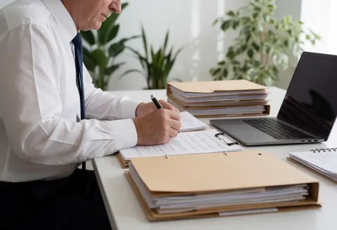 Senior lawyer reviewing documents and taking notes with a pen at a desk with a computer, folders, and plants in a clean professional setting.