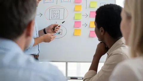 A group of professionals gathers around a whiteboard to review a diagram, with one person pointing to details while others observe and engage in the discussion.