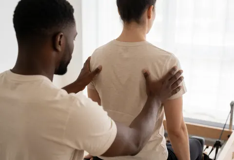 A therapist adjusts a person's posture while seated on a reformer in a physical therapy studio.