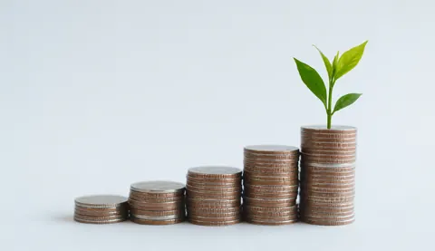 A stack of coins arranged in an ascending bar chart shape with a small green plant growing from the top coin, symbolizing financial growth and sustainable investment.