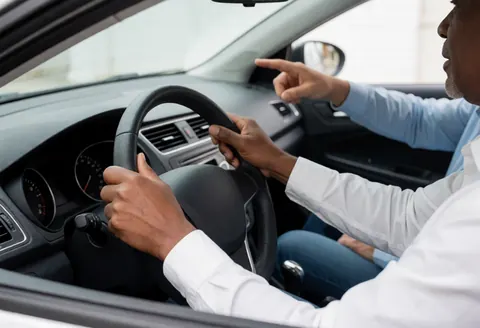 A driving instructor sits in the driver's seat with one hand on the steering wheel and the other pointing forward to guide a student during a lesson.