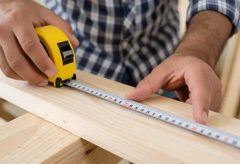A carpenter measures a wooden plank with a yellow tape measure in a workshop.