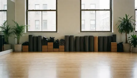 Empty yoga studio with wooden floors, yoga mats, and plants, lit by natural light from windows.