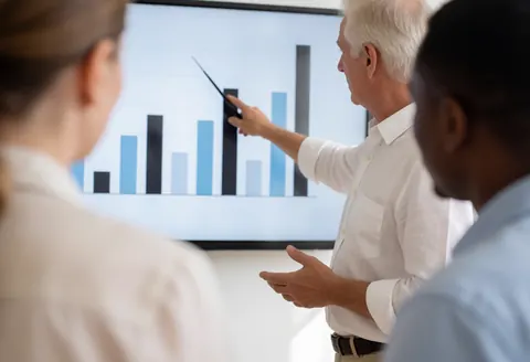A man in a white shirt points to a bar graph on a large screen while two people listen attentively in a meeting room.