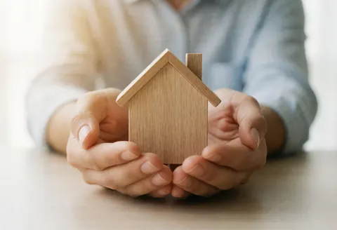 Two hands hold a wooden house model on a table, symbolizing protection and care for home.