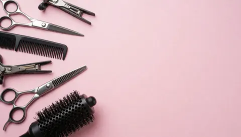 Hairdressing scissors, comb, and brush arranged on a light pink background, ideal for text and salon design