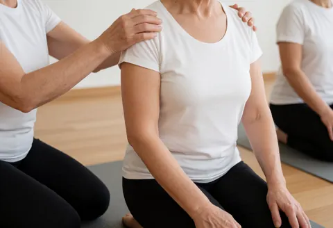 Yoga instructor adjusting a student's shoulder while kneeling in a studio with wooden floors and natural light