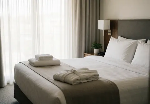 Clean hotel bed with white sheets, folded towels, and a robe, near a window with curtains.