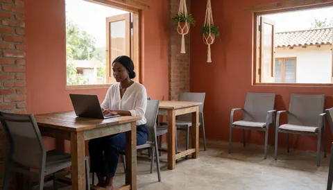 A woman works on a laptop at a wooden table in a coworking space with terracotta walls and natural light.
