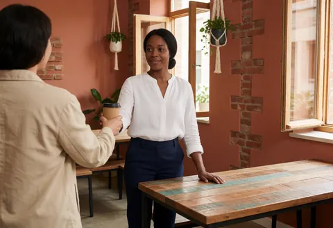 A staff member smiles while handing a coffee cup to a client in a coworking space with brick walls and wooden tables.