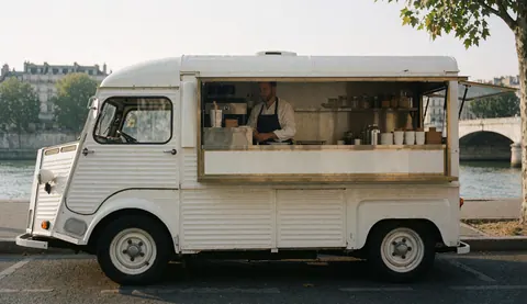 A white coffee truck is parked by the Seine river, with a man preparing drinks behind the counter.