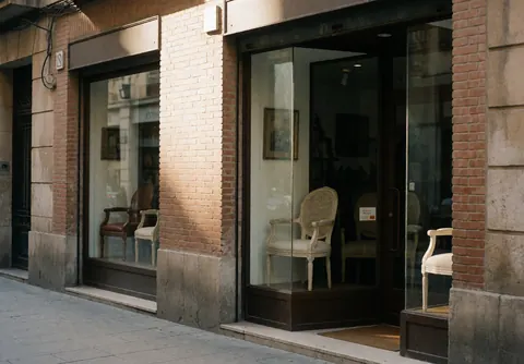 Facade of a furniture store with glass display windows showcasing classic style chairs and furniture on a street in Barcelona.