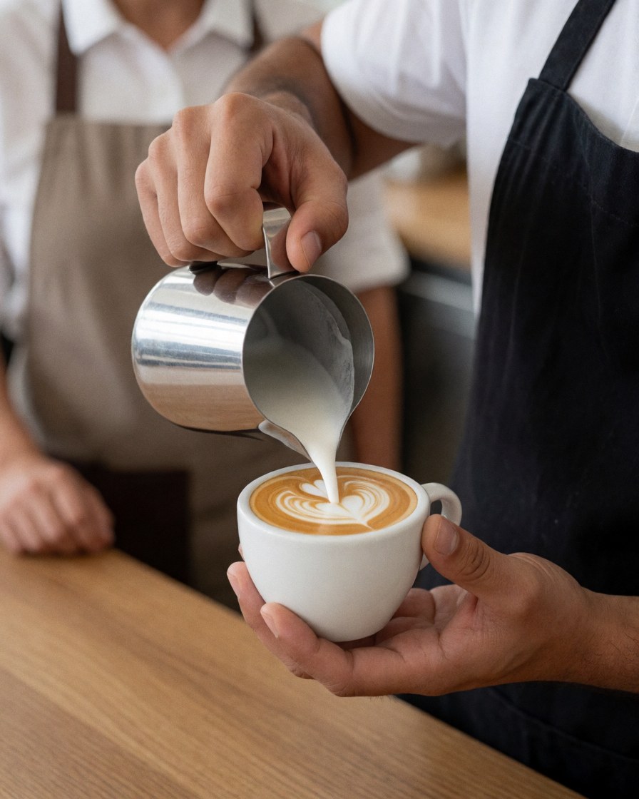 Barista in black apron pours milk into a cup of coffee with latte art on a wooden counter.