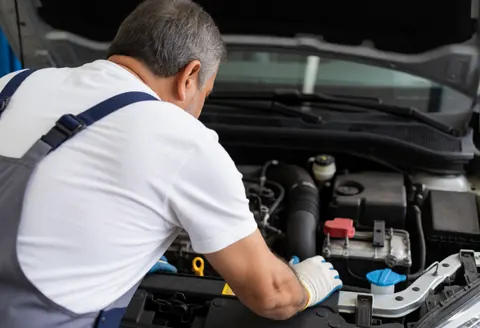 A mechanic wearing gloves inspects a car engine in a professional workshop with visible tools and components.