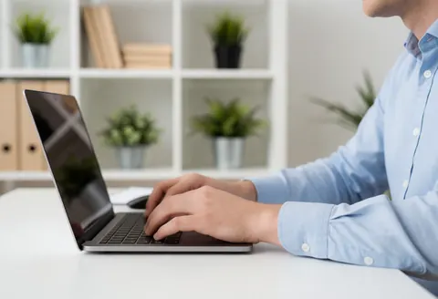 A man in a blue shirt types on a laptop on a white desk in an office with shelves and plants.
