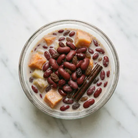 Glass of sweet beans dessert topped with cinnamon, raisins, and fruit on white marble surface