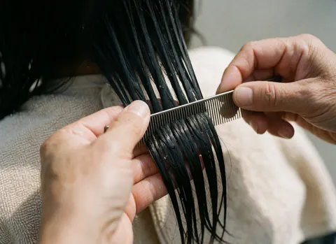 Close-up of hands using a fine-toothed comb on wet, shiny black hair with a beige towel in a professional salon setting