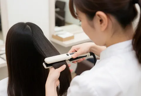 Professional stylist in white uniform using a ceramic flat iron to straighten long, shiny hair in a modern salon setting.