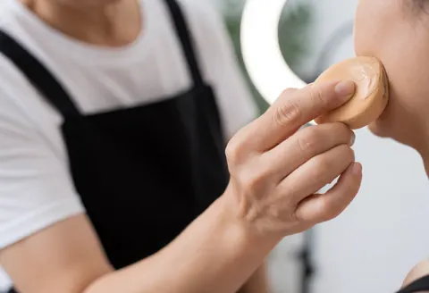 Professional makeup artist applying foundation with a damp sponge to a client's profile using ring light lighting in a blurred salon setting.