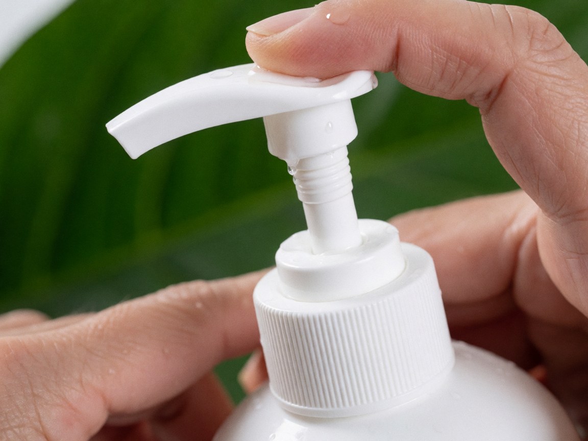 Close-up of a white shampoo pump bottle with water droplets, held by hands against a green leaf background, showcasing a fresh and natural hair care product.