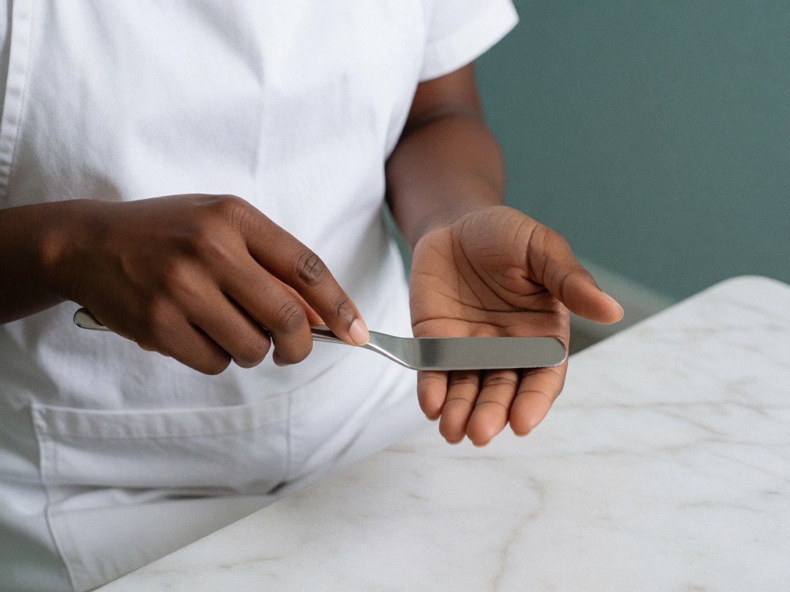 Aesthetician hands using a spatula to apply clear gel onto a palm in a clinic with white marble and turquoise walls