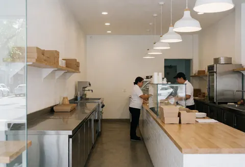 Bright and tidy taqueria interior featuring a white counter, stainless steel cooking equipment, wooden tables, and a clean minimalist design suitable for fast food businesses.