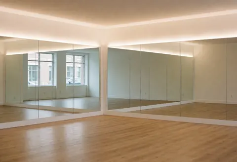 Spacious empty yoga studio featuring wall-to-wall mirrors, light wood flooring, and warm natural light creating a calm atmosphere for fitness classes.