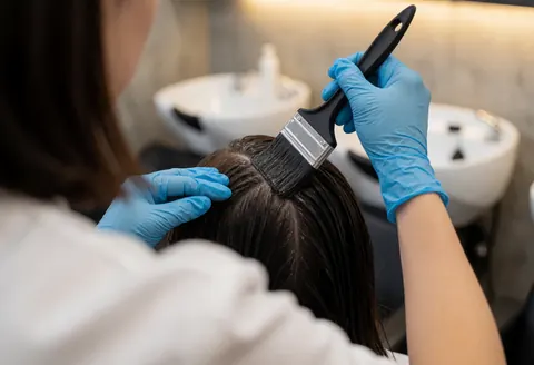 A stylist wearing blue gloves uses a brush to apply dark hair dye to a client's roots in a salon with white sinks and concrete walls.