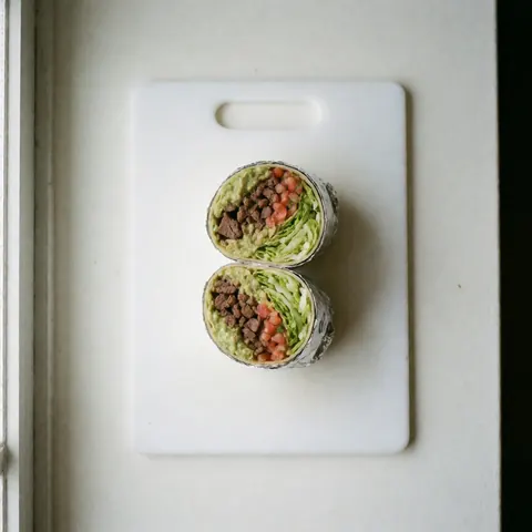 A halved burrito filled with meat, guacamole, lettuce, and pico de gallo resting on a white cutting board under natural light.