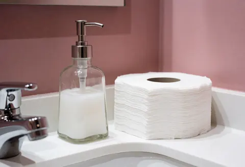 Bathroom vanity featuring a chrome faucet, soap dispenser, and paper towels against a pink wall, suitable for salon or beauty business decor