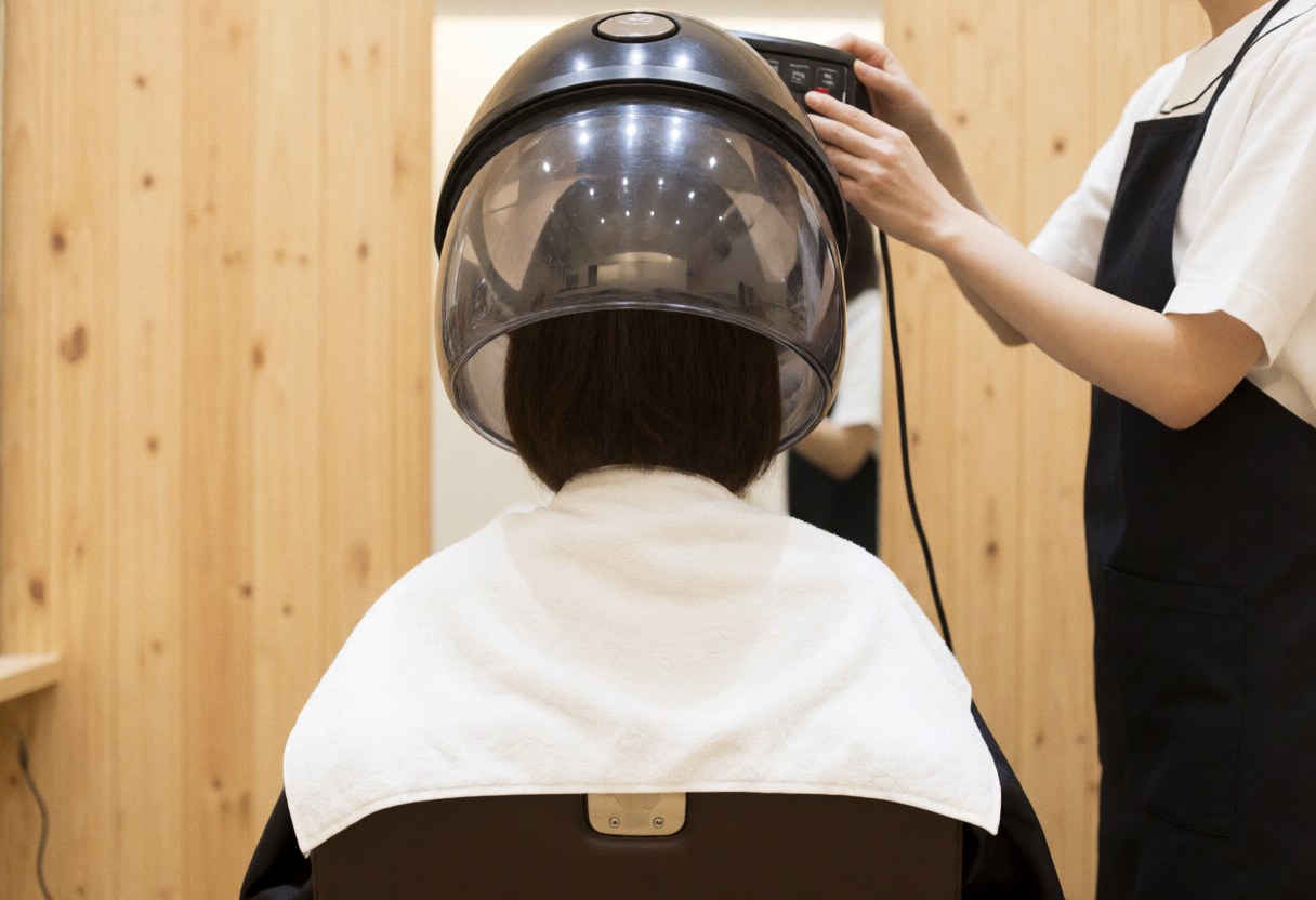Client with white towel under large hair dryer while stylist adjusts controls in wood-paneled salon