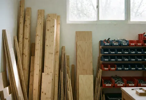 A tidy storage area filled with stacked wood planks, plywood sheets, and various tools near a window letting in natural light