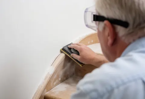 Back view of hands sanding a wooden ladder, creating dust, wearing safety glasses for protection against the white wall background