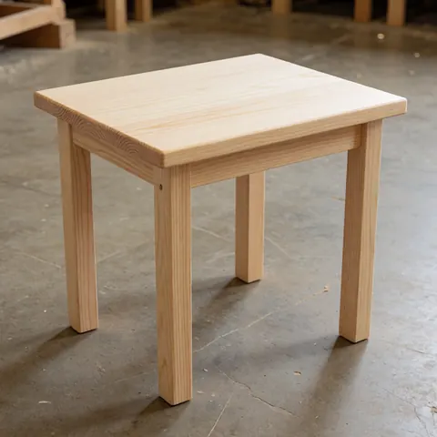 A simple furniture piece made from light radiata pine wood with visible grain texture, placed on a concrete floor in a workshop setting with natural lighting.