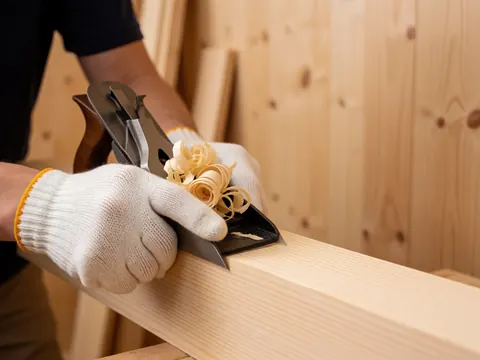 Close-up view of hands wearing protective gloves sanding a pine wood board, creating fine wood shavings and highlighting the natural wood texture during detailed craftsmanship work