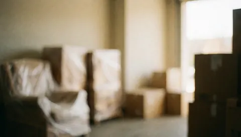 Out of focus warehouse scene featuring wrapped boxes and furniture illuminated by warm light, suitable for commercial text overlays and logistics services.