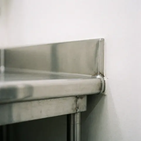 Close-up of polished stainless steel edge with visible weld on white wall, showing clean reflective surface ideal for professional food preparation areas