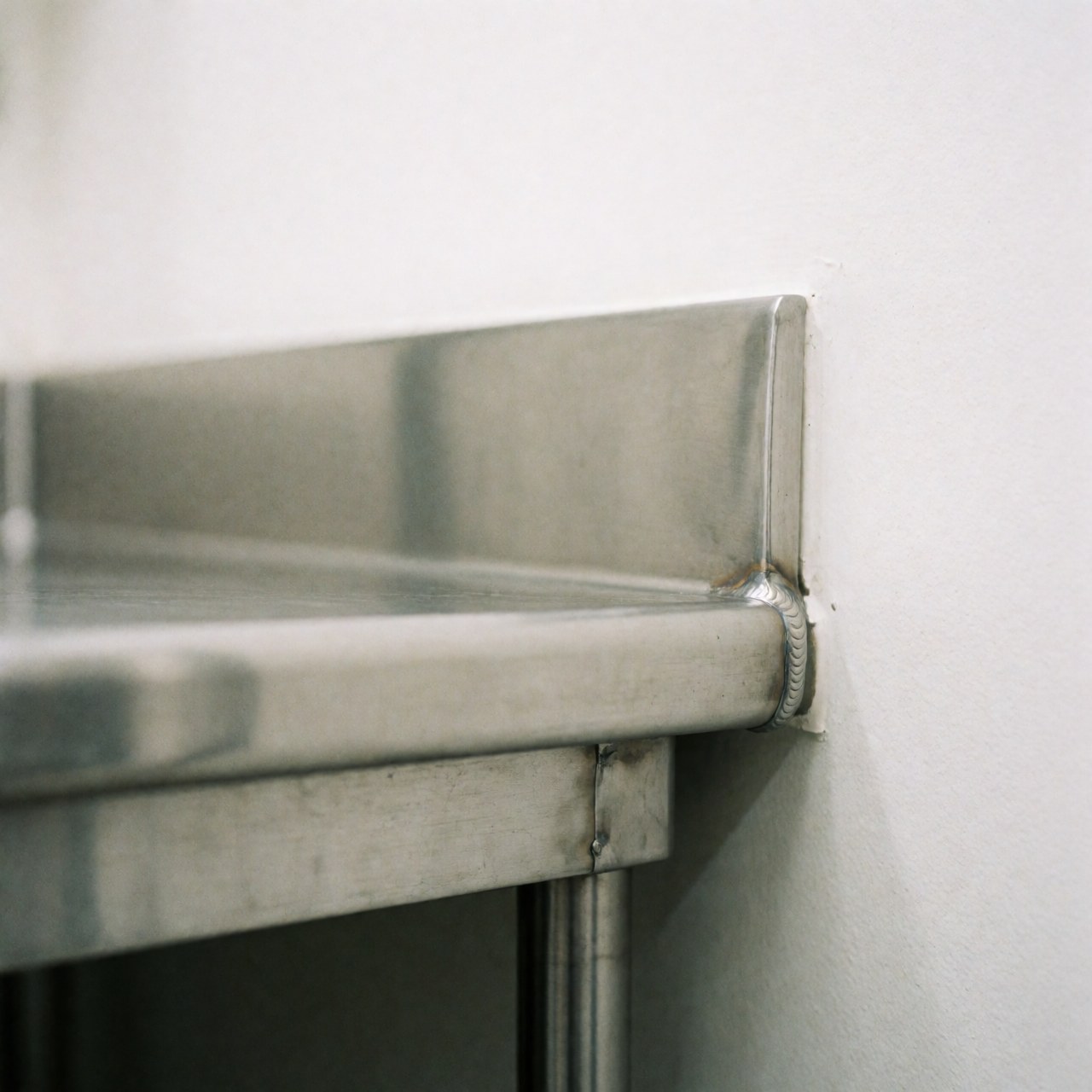 Close-up of polished stainless steel edge with visible weld on white wall, showing clean reflective surface ideal for professional food preparation areas