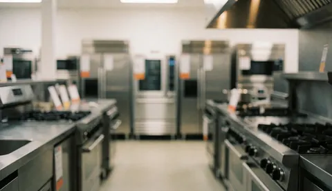 Clean showroom background featuring professional stainless steel kitchen appliances including stoves, ovens, refrigerators, and exhaust hoods, ideal for text overlay in marketing materials.