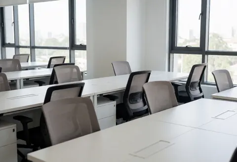 An empty modern coworking office interior featuring a clean white desk, ergonomic black chair, and large window with natural light and city view