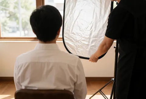 Photographer adjusts a light reflector while a client sits near a window for a professional corporate portrait session.