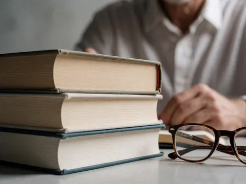 A pile of hardcover books with reading glasses resting on a work table against a concrete wall, creating an intellectual and professional atmosphere.