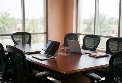 Empty corporate conference room featuring a wooden table, open laptops, black chairs, and natural light from windows creating a professional atmosphere.