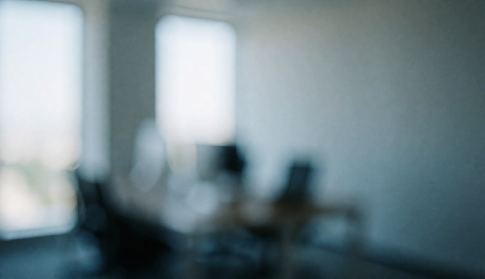 A clean and calm blurred office interior featuring white walls, blue and gray accents, a desk, and a chair near a window, suitable for text or advertising backgrounds.